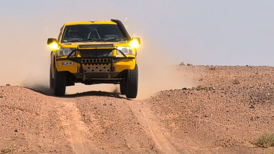 A yellow Land Rover desert racer driven by Pat Masters kick-starts sand on a vast, sun-drenched dune.