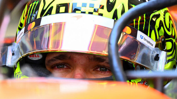 A close-up of a race driver wearing a bright neon yellow and black helmet with a clear visor, seated inside an orange race car cockpit.