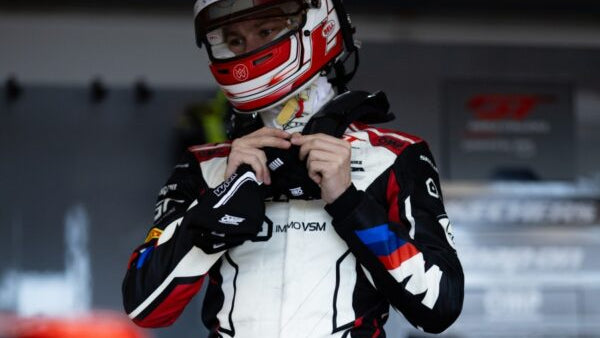Racing driver in a white and black suit with red accents, standing in a garage.